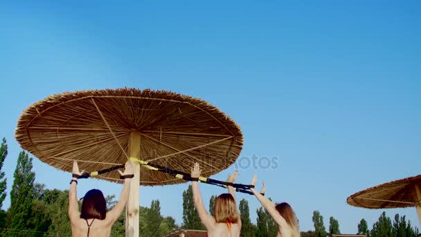 Três mulheres jovens atléticas, sensuais em maiôs, instrutores, fazendo exercícios com sistema de fitness trx, tiras de suspensão TRX. Na praia, no verão, ao pôr do sol, aos raios de sol . — Vídeo de Stock