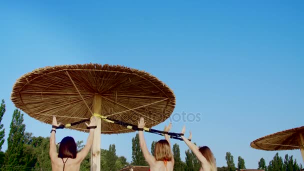 Drei athletische, sexy junge Frauen in Badeanzügen, Instruktoren, die Übungen mit Fitness-Trx-System, Trx-Federbeinen machen. am Strand, im Sommer, bei Sonnenuntergang, in der Sonne. — Stockvideo