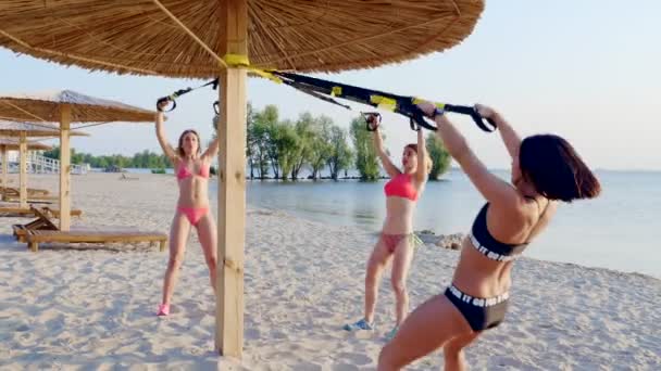 Three athletic, sexy young women in swimsuits, instructors, doing exercises with fitness trx system, TRX suspension straps. On the beach, in summer, at sunset, in sun rays. — Stock Video