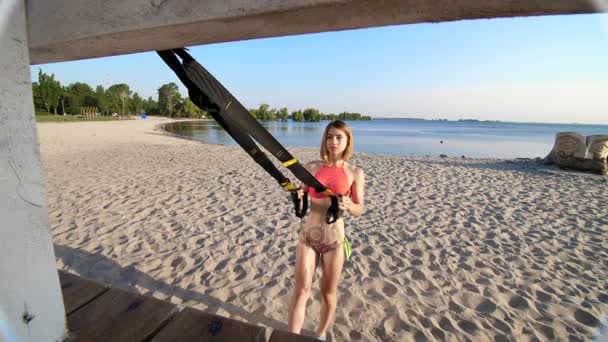 Athletic, sexy young woman in swimsuit, coach, instructor, performs, doing exercises with fitness trx system, TRX suspension straps. On the beach, summer, sunset, in sun rays. — Stock Video