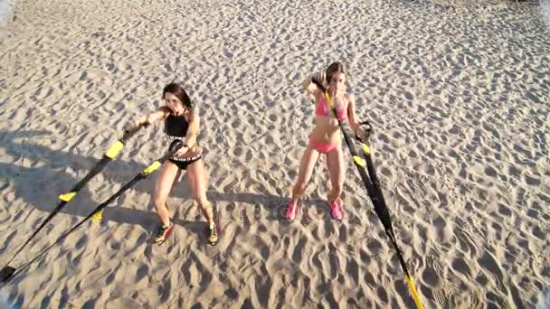 Two athletic, sexy young women in swimsuits, instructors, doing exercises with fitness trx system, TRX suspension straps. On the beach, in summer, at sunset, in sun rays. — Stock Video