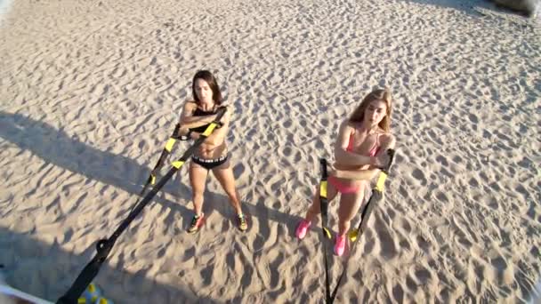 Две атлетичные, сексуальные девушки в купальниках, инструкторы, занимающиеся упражнениями с системой фитнес-трекс, ремни для подвески TRX. На пляже, летом, на закате, в солнечных лучах . — стоковое видео