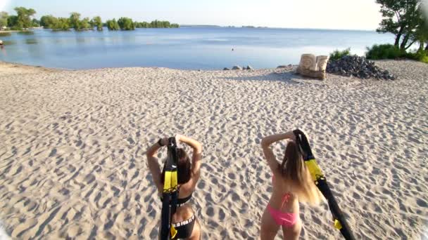 Due giovani donne atletiche e sexy in costume da bagno, istruttori, facendo esercizi con sistema fitness trx, cinghie di sospensione TRX. In spiaggia, in estate, al tramonto, sotto i raggi del sole . — Video Stock