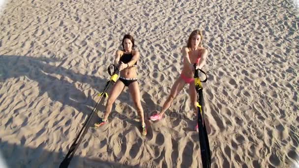 Dvě sportovní, sexy mladé ženy v plavkách, instruktoři, dělat cvičení s fitness systém trx, Trx popruhy suspenze. Na pláži, v létě, při západu slunce, sluneční paprsky. — Stock video