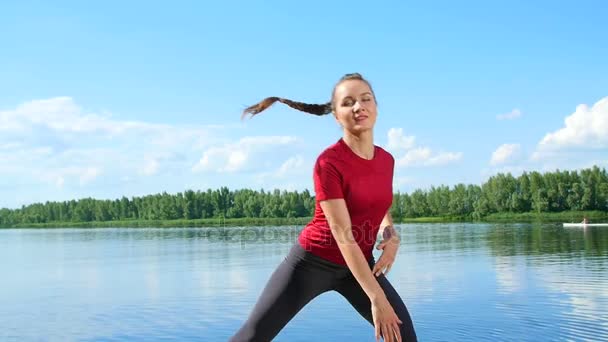 Schöne, athletische junge blonde Frau, die verschiedene Übungen macht. See, Fluss, blauer Himmel und Wald im Hintergrund, sonniger Sommertag. — Stockvideo