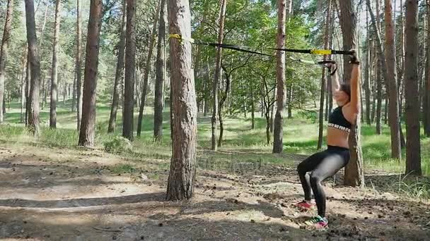 Hermosa, atlética, mujer joven sexy, entrenador, instructor, realiza ejercicios, haciendo ejercicios con sistema de trx fitness, correas de suspensión TRX. En el bosque de pinos, en verano, en los rayos del sol . — Vídeos de Stock