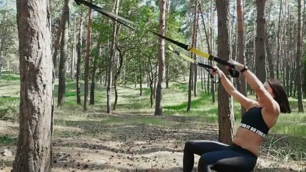 Hermosa, atlética, mujer joven sexy, entrenador, instructor, realiza ejercicios, haciendo ejercicios con sistema de trx fitness, correas de suspensión TRX. En el bosque de pinos, en verano, en los rayos del sol . — Vídeos de Stock