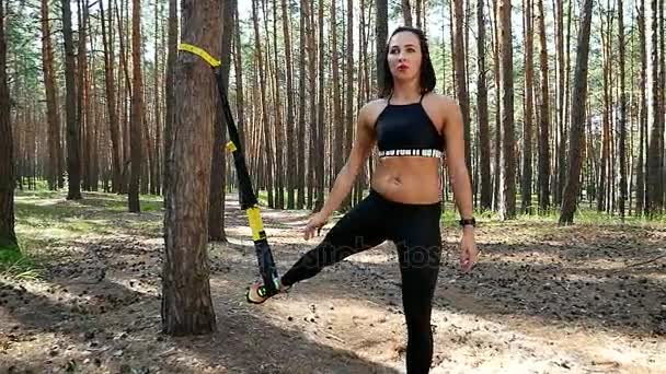 Hermosa, atlética, mujer joven sexy, entrenador, instructor, realiza ejercicios, haciendo ejercicios con sistema de trx fitness, correas de suspensión TRX. En el bosque de pinos, en verano, en los rayos del sol . — Vídeo de stock