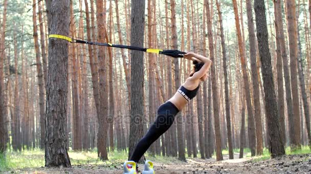 Mulher bonita, atlética, sexy jovem, treinador, instrutor, realiza exercícios, fazendo exercícios com sistema de fitness trx, correias de suspensão TRX. Na floresta de pinheiros, no verão, nos raios de sol . — Vídeo de Stock