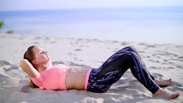 Здорова, молода красива жінка виконує вправи для м'язів преси, піднімає тіло, накачує пресу лежачи на піску, на пляжі, на сході сонця — стокове відео