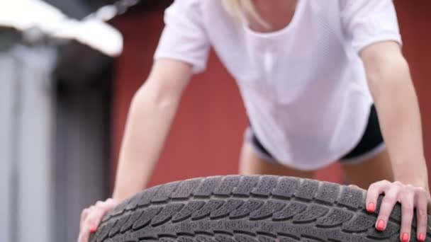 Sexy sportive jeune femme blonde en short, effectue divers exercices de force à l'aide de pneus, push-ups, En été, près de vieux garages abandonnés. gros plan — Video