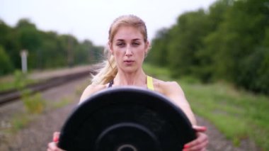 Güzel seksi atletik genç sarışın kadın siyah tozluk egzersizleri bir ağır siklet plakalı demiryoluna raylar, yaz aylarında performans. Ağır çekim. portre