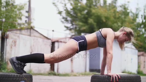 Sexig atletisk ung blond kvinna i shorts, utför olika styrkeövningar med hjälp av däck, armhävningar, på sommaren, nära gamla övergivna garage — Stockvideo