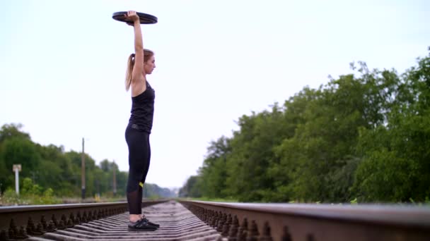 Mooie sexy atletische jonge blonde vrouw in zwarte legging uitvoeren van oefeningen met een zwaar gewicht plaat, op de spoorweg, op de rails, dwarsliggers, in de zomer. Slow motion — Stockvideo