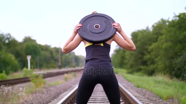 Schöne sexy, athletische junge blonde Frau in schwarzen Leggings, die im Sommer Übungen mit einer schweren Hantelscheibe macht, auf der Schiene, auf Schienen, auf Schwellen. Zeitlupe — Stockvideo
