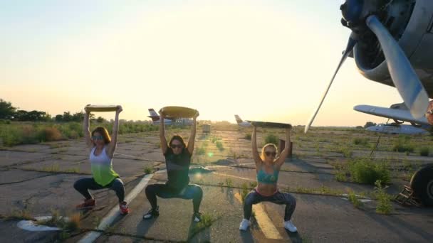 Ao pôr-do-sol, atléticas, jovens mulheres em óculos de sol, em collants, fazendo exercícios com peso, placa de peso pesado, em um aeródromo abandonado, contra o fundo de um avião velho , — Vídeo de Stock