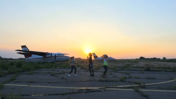 Belles, athlétiques, jeunes femmes en lunettes de soleil, en collants, effectuer synchroniquement différents exercices de force, sauts, sit-ups. sur un aérodrome abandonné, près de l'avion, au coucher du soleil . — Video