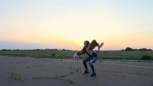 Hermosa, atlética, joven en gafas de sol, en mallas, realiza sentadillas, sosteniendo a una chica en sus brazos, ejercicios en parejas, como un peso adicional. en un aeródromo abandonado, puesta de sol — Vídeo de stock