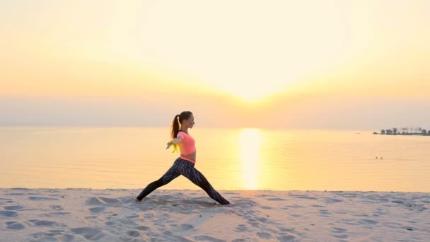 Здоровая, молодая красивая женщина медитирует, растягивает, практикует йогу на морском пляже, на восходе солнца, делает упражнения для равновесия и координации, глубокий мышечный тонус — стоковое видео