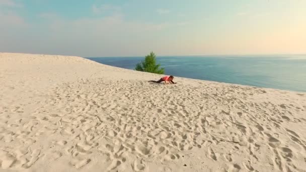 Mujer joven y saludable en gafas de sol meditando, estirando, practicando yoga en la playa del mar, al amanecer, hace ejercicios para el equilibrio, coordinación, tono muscular profundo . — Vídeos de Stock