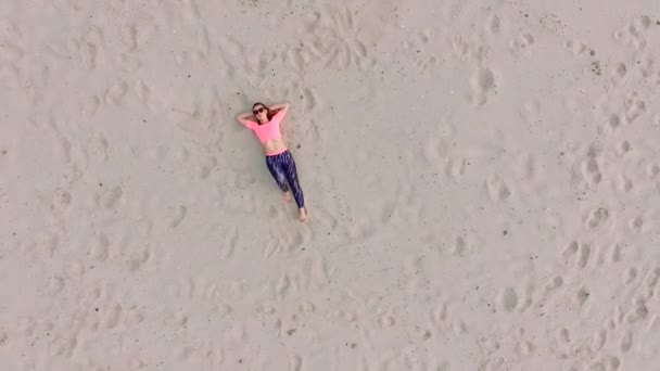 Κομψό νεαρό αθλητική όμορφη γυναίκα σε γυαλιά ηλίου, ξαπλωμένοι στην άμμο, σε μια έρημη παραλία, καλοκαίρι. Θέα από ψηλά, aero βίντεο. — Αρχείο Βίντεο