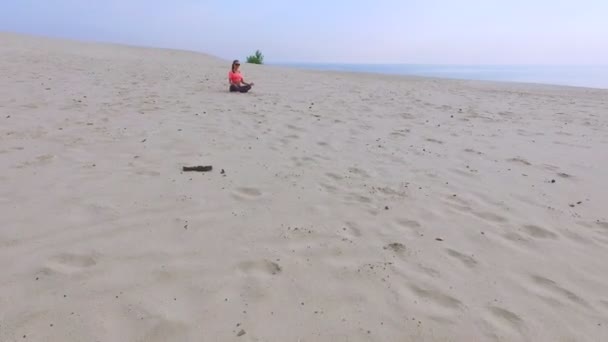 Elegante joven hermosa mujer en gafas de sol, sentada en la arena en una pose de loto, meditando, practicando yoga en una playa desierta, en verano. Vista desde arriba, aero video — Vídeo de stock