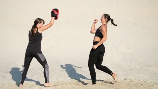 Две атлетичные, молодые женщины в черных костюмах занимаются парой, тренируются ногами, на пустынном пляже, под голубым небом, летом, под жарким солнцем. Медленное движение — стоковое видео