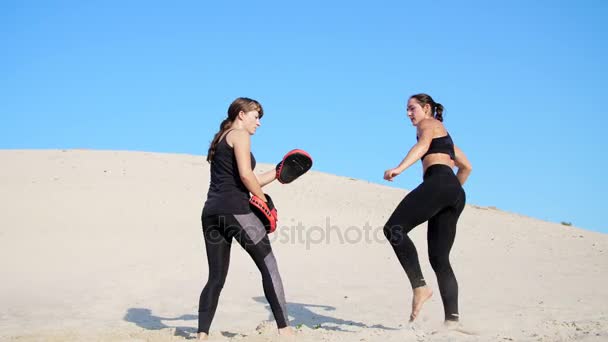 Deux jeunes femmes sportives en costume de fitness noir sont engagées dans une paire, accomplissent des coups de pied, s'entraînent à se battre, sur une plage déserte, contre un ciel bleu, en été, sous un soleil chaud. Mouvement lent — Video
