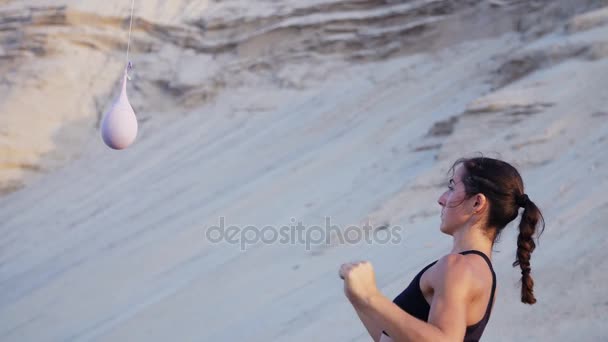 Chica atlética en un top negro, práctica patadas, patadas pies delante del globo. Chica patea globo con su pie, y derrames de agua de colores, espolvorea de ella, En una playa desierta, en verano . — Vídeo de stock