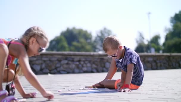 Children, a boy and a girl in sunglasses, paint with colored crayons on the asphalt, street tiles. A hot summer day. — Stock Video