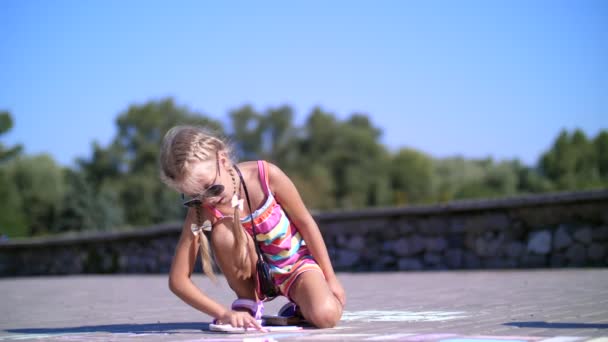 Een meisje in zonnebril, trekt tekeningen met gekleurde krijtjes op het asfalt, straat tegels. Een warme zomerdag. — Stockvideo