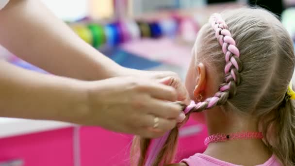 Vacker blond flicka, sju år gammal, flätad två flätor, göra en frisyr med rosa hårlockar i en skönhetssalong, en salong med frisörer, framför en stor spegel. en liten prinsessa — Stockvideo