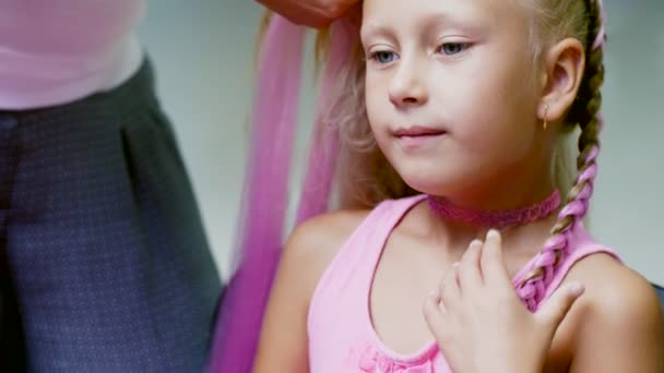 Hermosa chica rubia, de siete años, trenzado dos coletas, hacer un peinado con mechones de pelo de color rosa en un salón de belleza, un salón de peluquería, frente a un gran espejo. una princesita — Vídeo de stock