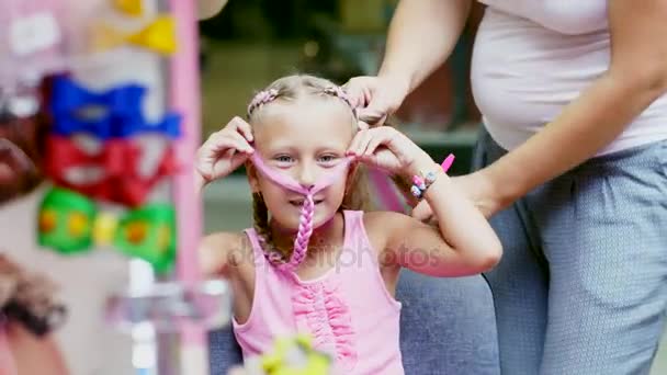 Belle fille blonde, de sept ans, tressée de deux nattes, faire une coiffure avec des mèches roses de cheveux dans un salon de beauté, un salon de coiffure, devant un grand miroir. une petite princesse — Video