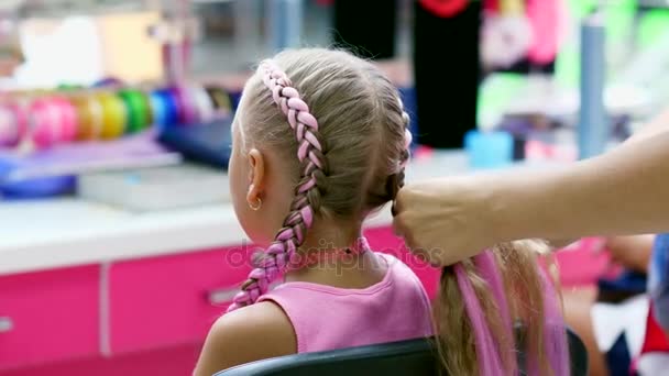 Hermosa chica rubia, de siete años, trenzado dos coletas, hacer un peinado con mechones de pelo de color rosa en un salón de belleza, un salón de peluquería, frente a un gran espejo. una princesita — Vídeo de stock