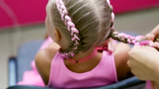 Vacker blond flicka, sju år gammal, flätad två flätor, göra en frisyr med rosa hårlockar i en skönhetssalong, en salong med frisörer, framför en stor spegel. en liten prinsessa — Stockvideo