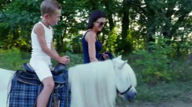 Bir kadın ve bir çocuk etrafında alanı yürüyor, oğlum bir midilli sürme, anne midilli a at başlığı için bekletme. Neşeli, mutlu aile tatili. Forest yakınındaki yaz aylarında açık havada