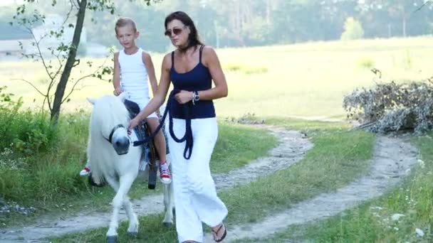 Seorang wanita dan anak laki-laki berjalan di sekitar lapangan, anak mengendarai kuda poni, ibu memegang kuda poni untuk kekang. Liburan keluarga bahagia dan ceria. Di luar ruangan, di musim panas, dekat hutan — Stok Video