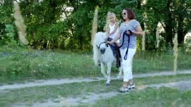Anne ve kızı alanı yürüyor, kızı bir midilli sürme, anne midilli a at başlığı için bekletme. Neşeli, mutlu aile tatili. Forest yakınındaki yaz aylarında açık havada