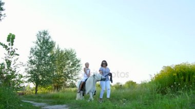 Anne ve kızı alanı yürüyor, kızı bir midilli sürme, anne midilli a at başlığı için bekletme. Neşeli, mutlu aile tatili. Forest yakınındaki yaz aylarında açık havada