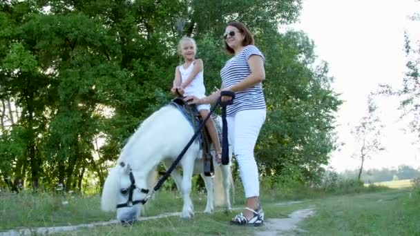 Ibu dan anak perempuan berjalan di sekitar lapangan, anak perempuan naik kuda poni, ibu memegang kuda poni untuk kekang. Liburan keluarga bahagia dan ceria. Di luar ruangan, di musim panas, dekat hutan — Stok Video