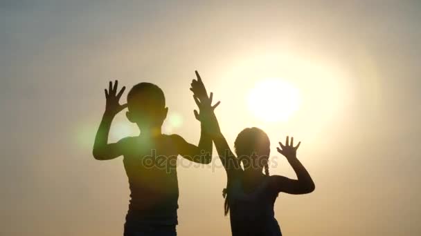Silhuetas, figuras de crianças, menino e menina pulando, se divertindo, dançando, abraçando no fundo do sol, ao pôr do sol no verão. Família feliz. Movimento lento — Vídeo de Stock