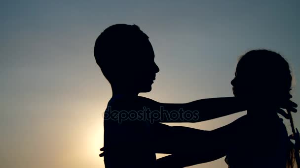 Силуэты, фигуры детей, мальчика и девочки, брат и сестра, обнимающиеся на фоне солнца, на закате, летом — стоковое видео
