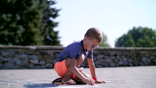 A boy draws drawings with colored crayons on the asphalt, street tiles. A hot summer day. — Stock Video