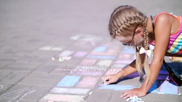 Uma menina em óculos de sol, desenha desenhos com lápis de cor no asfalto, azulejos da rua. Um dia quente de verão . — Vídeo de Stock