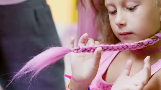 Menina loira bonita, de sete anos de idade, trançou duas tranças, fazer um penteado com fechaduras cor-de-rosa de cabelo em um salão de beleza, um salão de cabeleireiro, na frente de um grande espelho. uma pequena princesa — Vídeo de Stock