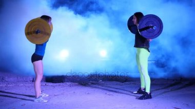 İki atletik kızlar, sporcular, halter yükseltmek, mekik halter ile yap. Geceleri, projektörler, hangarda bir eski terk edilmiş, Bina hafif duman, sis ışığında.