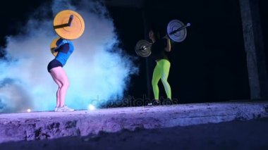 İki atletik kızlar, sporcular, halter yükseltmek, mekik halter ile yap. Geceleri, projektörler, hangarda bir eski terk edilmiş, Bina hafif duman, sis ışığında.