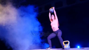 Ağırlıkları ile çeşitli güç egzersizleri yapıyor atletik, güzel genç bir kadın. Geceleri, projektörler, hangarda bir eski terk edilmiş, Bina hafif duman, sis ışığında