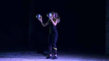 Ağırlıkları ile çeşitli güç egzersizleri yapıyor atletik, güzel genç bir kadın. Gece, terk edilmiş eski bir hangarda hafif duman, siste çok renkli projektörler ışığında,